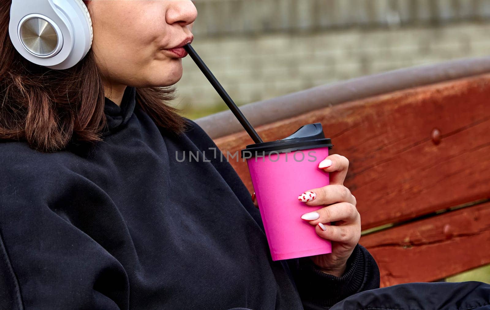 A young woman with long brown hair and white headphones is drinking from a pink cup with a straw. She is wearing a black sweatshirt and has her nails painted pink with white polka dots.