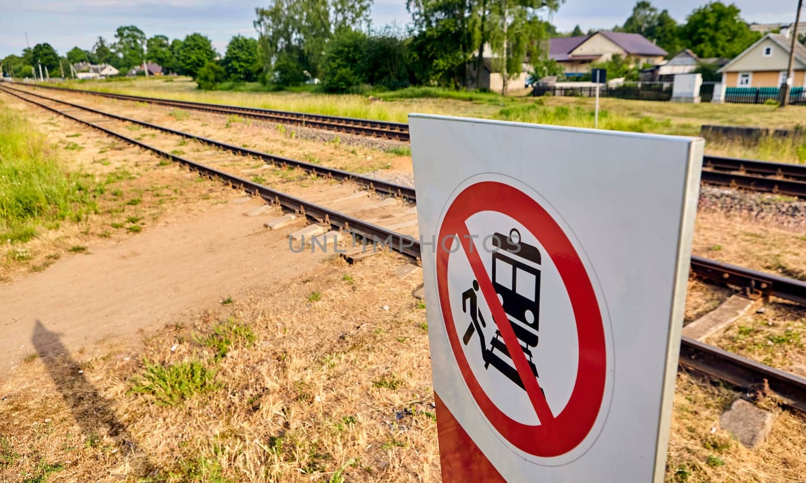 A concrete railway track under a sunny sky with a vivid prohibition sign displaying a train and a person, emphasizing no crossing. The scene captures rail safety on a lovely day.