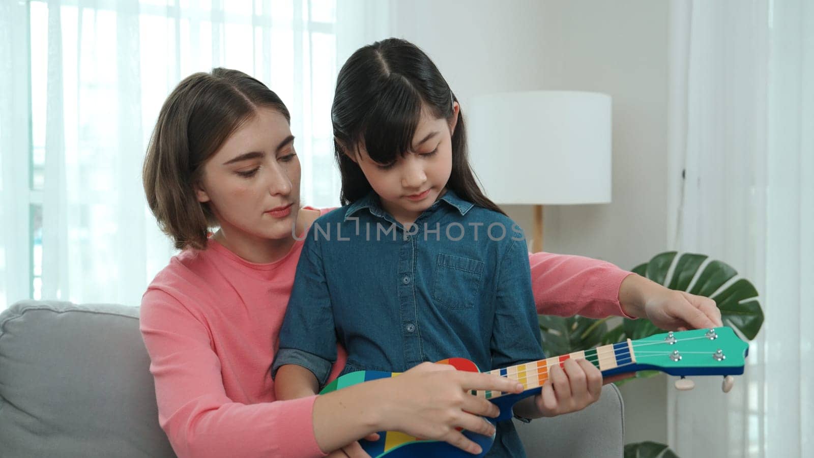 Happy girl playing ukulele while caucasian mom teaching and explain about acoustic music at home. Cute child learning about instrument. Happy caucasian mother and girl spend time together. Pedagogy.