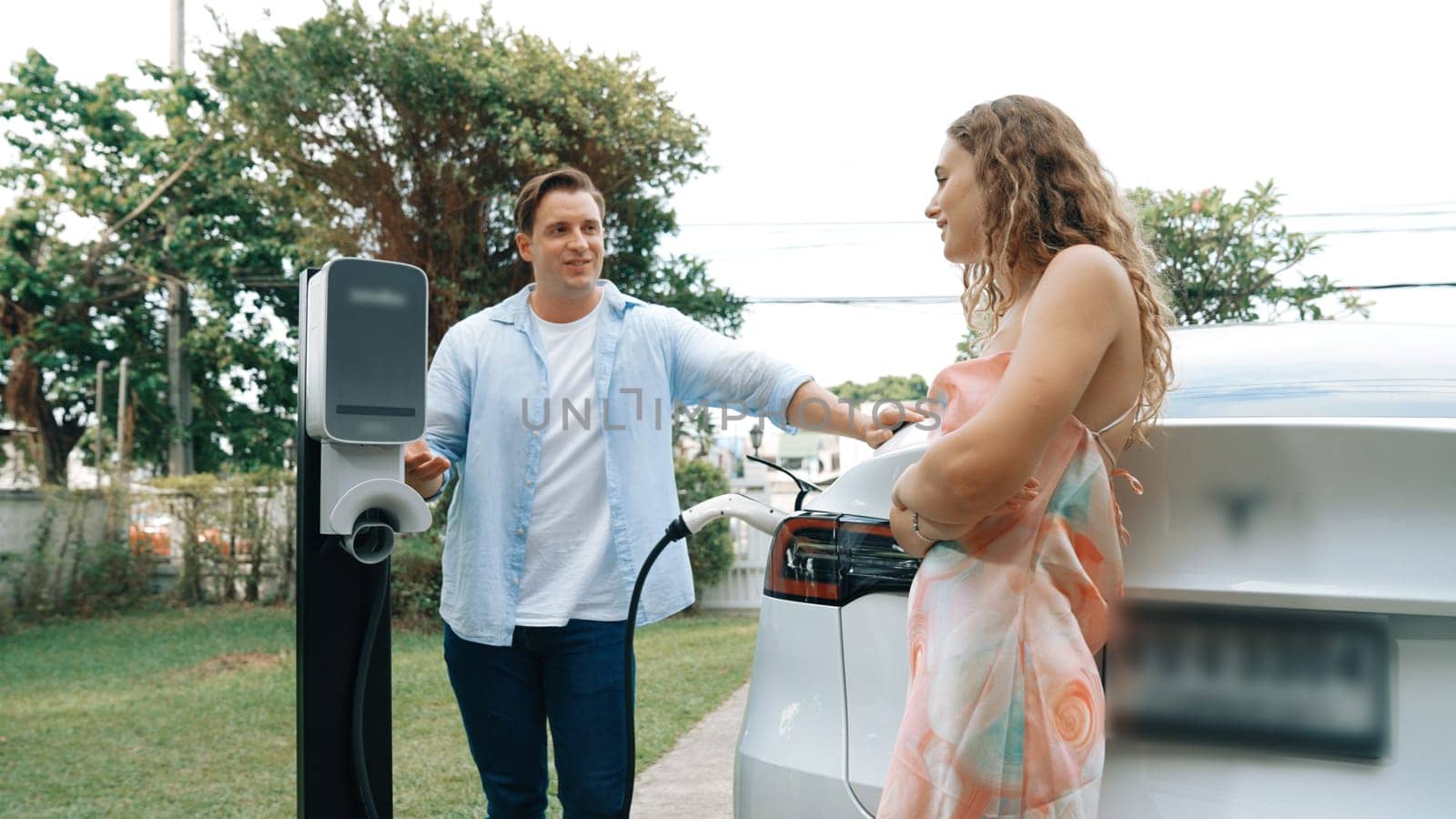 Happy and lovely couple with eco-friendly conscious recharging electric vehicle from EV charging station. EV car technology utilized as alternative transportation for future sustainability. Synchronos