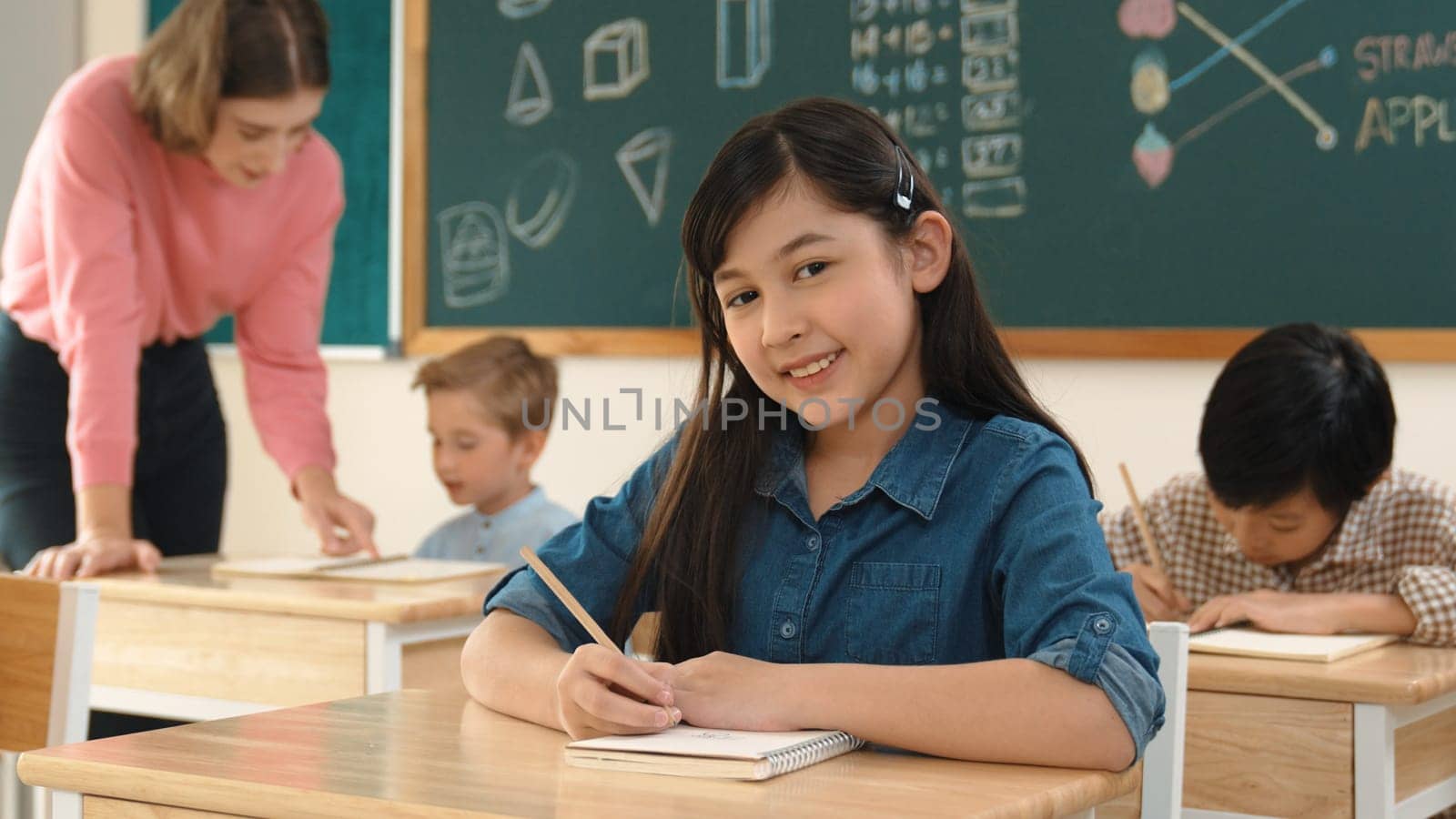 Asian girl writing test or classwork while looking at camera at classroom. Diverse happy student taking notes and teacher help caucasian boy doing test while sitting in front of blackboard. Pedagogy.