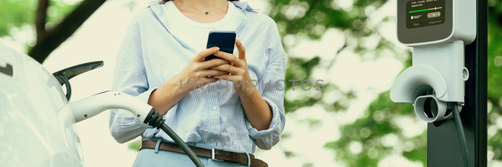 Young woman using smartphone online banking application to pay for electric car battery charging from EV charging station during vacation road trip at national park or summer forest. Panorama Exalt