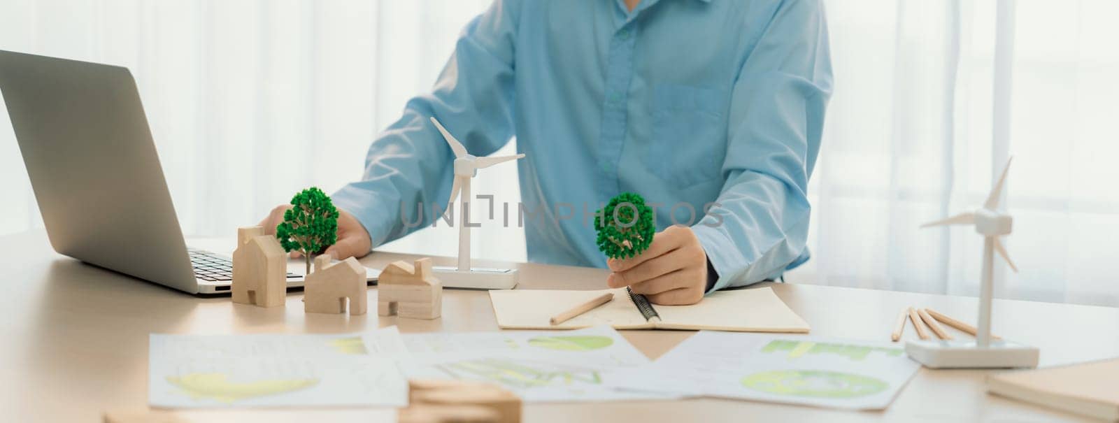 Professional architect plans to use green design in an eco house. Skilled engineer design an eco house blueprint by using laptop on table with house and windmill model scatter around. Delineation.