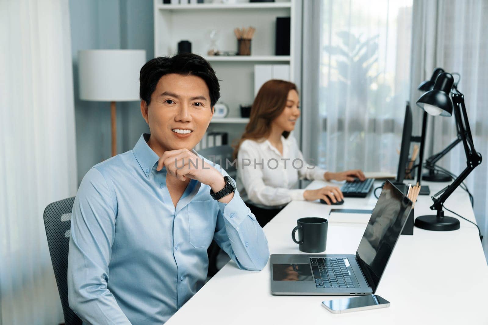 Profile smiling of smart Asian businessman looking camera to pose at modern office. Blurry background woman colleague working on pc to check email marketing plan analysis report at desk. Infobahn.