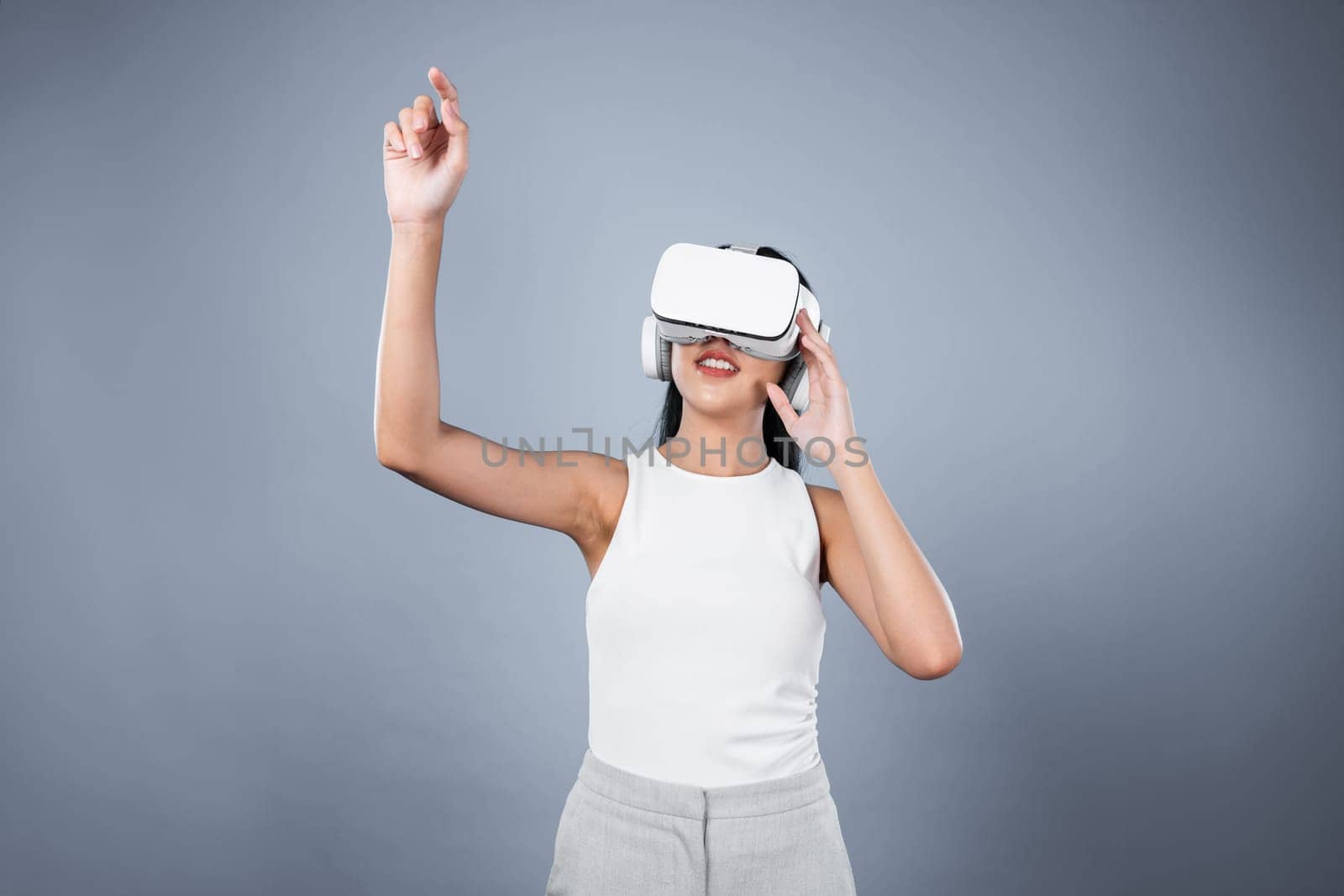 Smart female standing with gray background wearing VR headset connecting metaverse, futuristic cyberspace community technology. Elegant woman excited seeing generated virtual scenery. Hallucination.