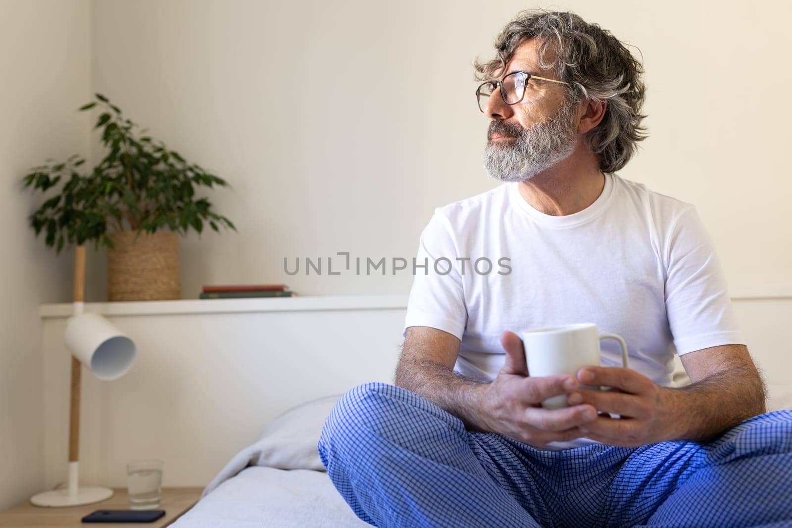 Pensive mature man relaxing, sitting on bed enjoying morning coffee looking away. Copy space. Lifestyle concept.