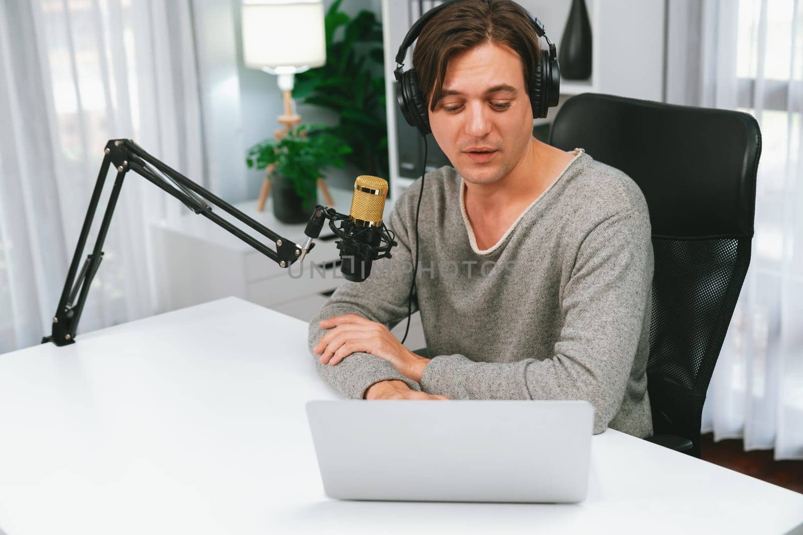 Host channel in smiling smart broadcaster recording, talking show with script and smartphone on desk on live social media, wearing headphones to record video streamer at modern home studio. Pecuniary.