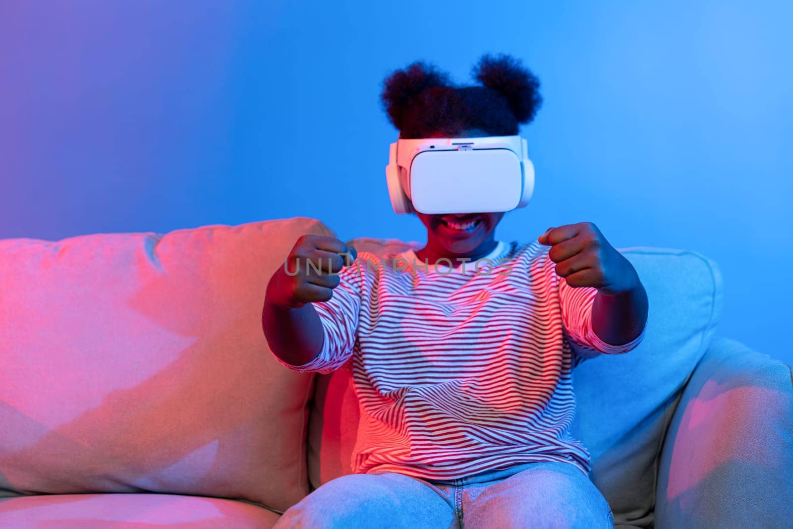 Smiling African American woman driver wearing VR playing car racing turning metaverse sitting on living sofa at vibrant blue pink neon light gaming technology 3D graphic car competition. Contrivance.
