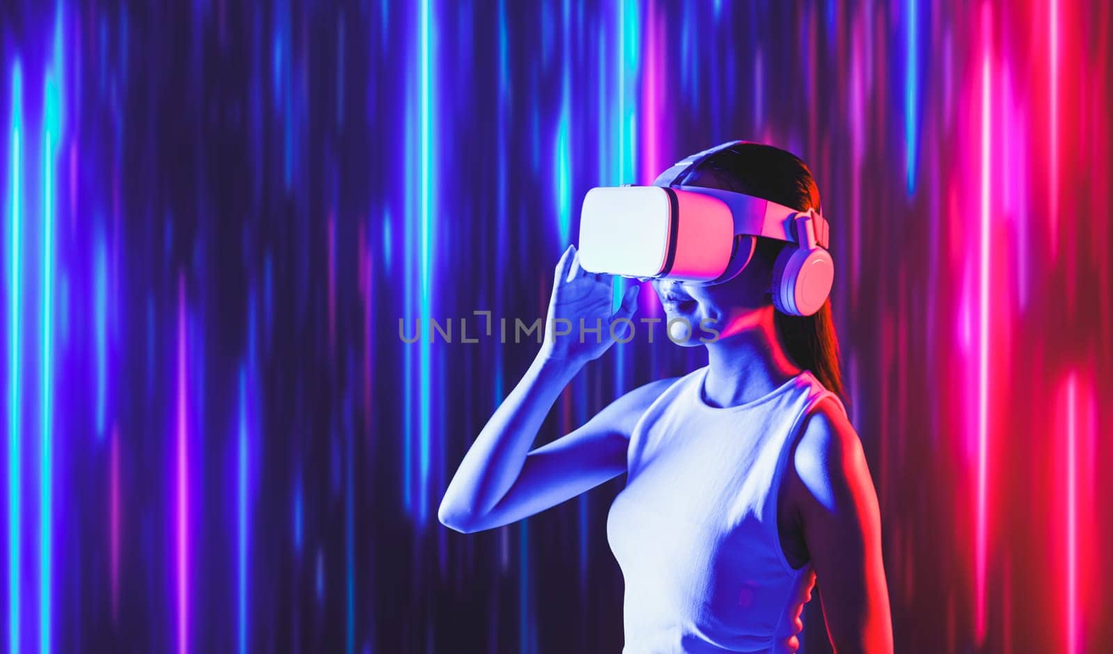 Smart female standing surrounded by neon light wearing VR headset connecting metaverse, future cyberspace community technology. Elegant woman looking faraway and smiling satisfactorily. Hallucination.