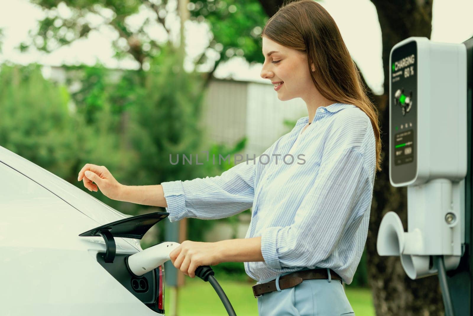 Young woman recharge EV electric vehicle's battery from EV charging station in outdoor green city park scenic. Eco friendly urban transport and commute with eco friendly EV car travel. Exalt