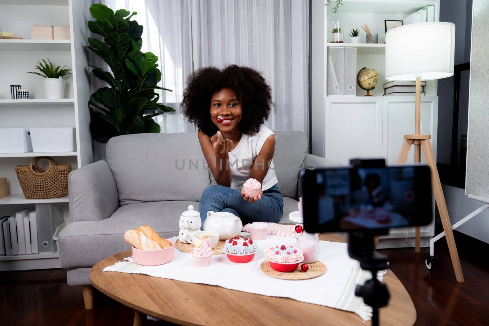 Beautiful young African blogger presenting piece of cupcake in concept special cuisine with video record on smartphone. Content creating of social media with favorite sweets bakery dish. Tastemaker.