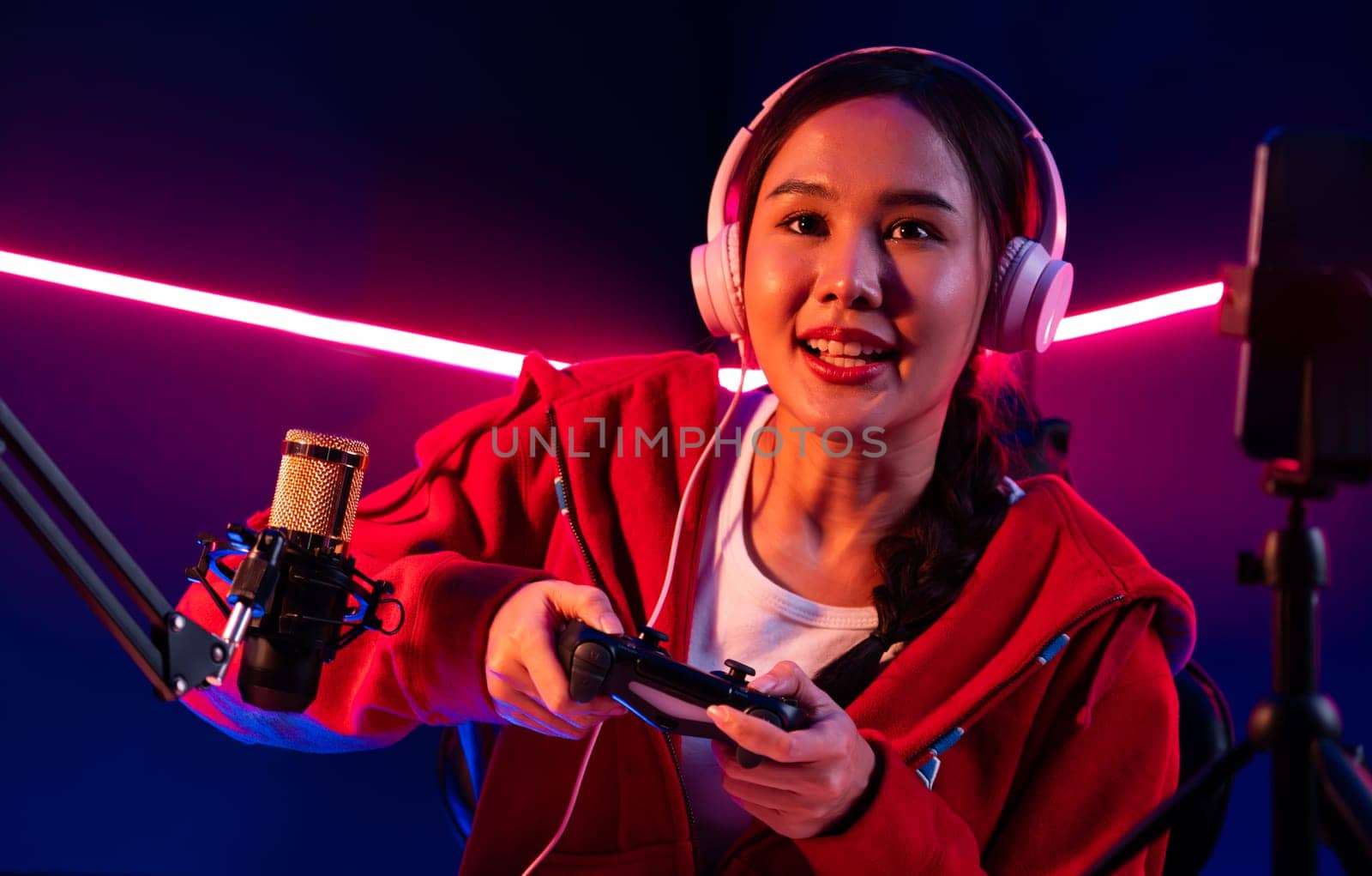 Host channel of smiling beautiful Asian girl streamer with joystick playing online game wearing headphones paste talking with viewers media online. Esport skilled team players in neon room. Stratagem.