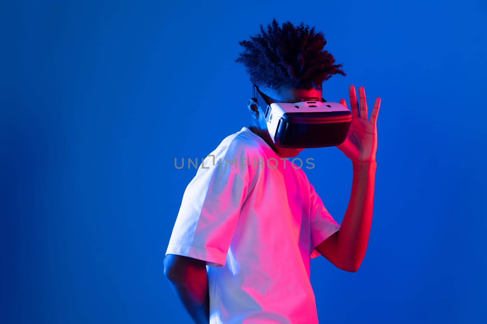 Surprised young African American looking through VR holding interesting object hologram on pink blue neon wall at metaverse world connecting digital futuristic technology virtual reality. Contrivance.