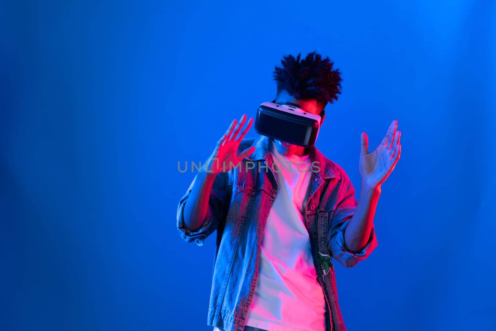 Smiling young African American looking through VR dancing in song with hologram gaming on pink blue neon wall at metaverse world connecting digital futuristic technology virtual reality. Contrivance.