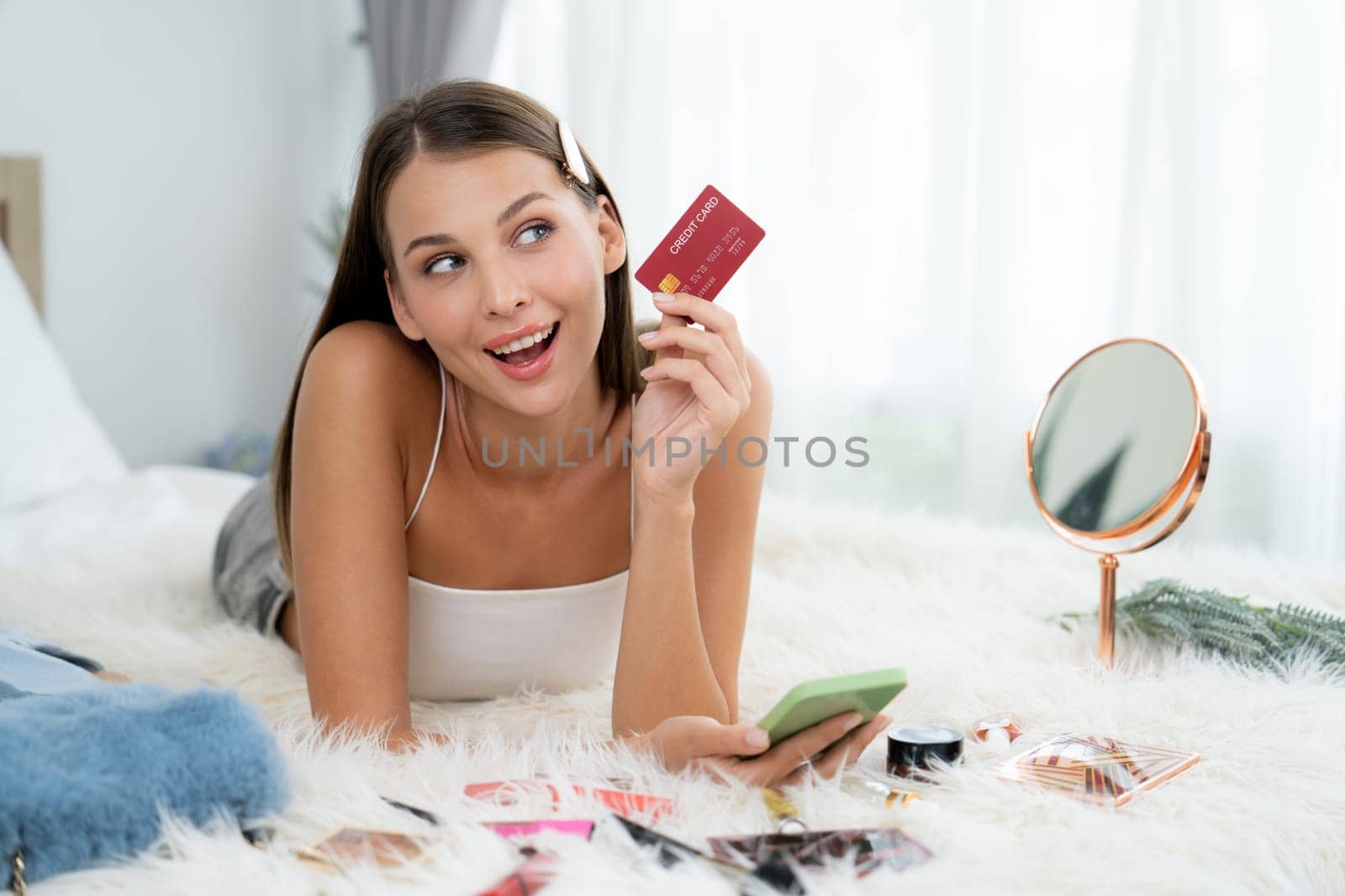 Young woman lying on the bed using online payment app and digital wallet on smartphone to pay with credit card. E commerce shopping and modern purchasing via mobile internet. Unveiling