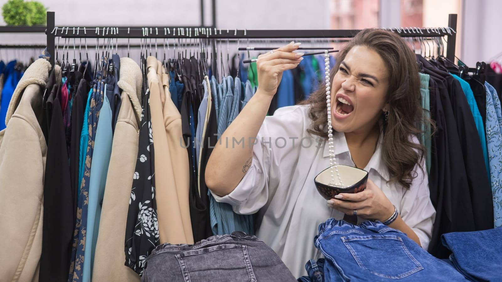 Happy fat woman fooling around and pretending to eat a pearl necklace with Chinese chopsticks in a clothing store for large people