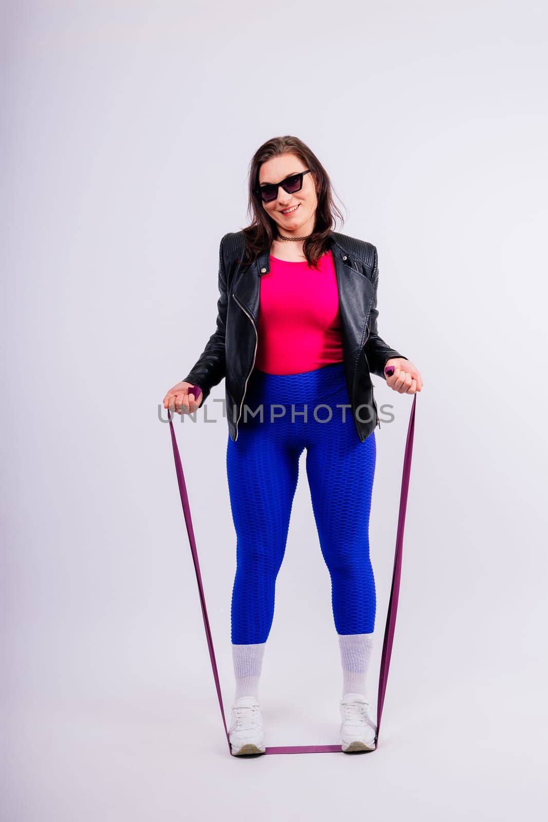 Strong female working with resistance band. Photo of model in fashionable sportswear on grey background.