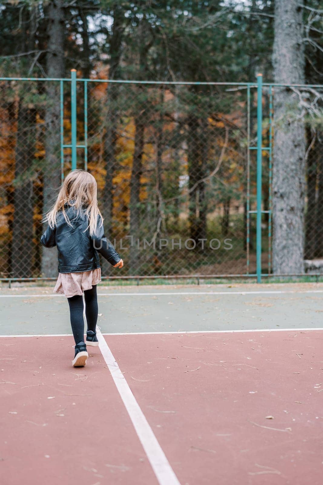 Little girl walks along the sports ground towards the mesh fence. Back view. High quality photo