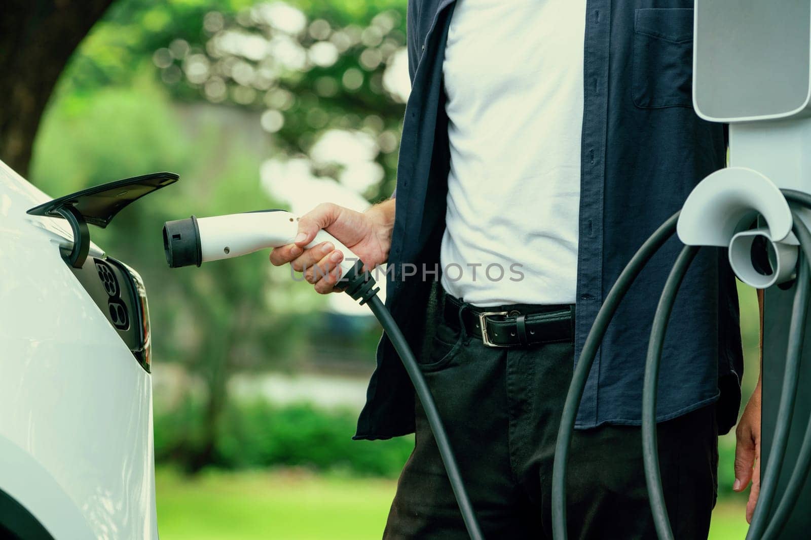 Man recharging battery for electric car during road trip travel EV car in natural forest or national park. Eco friendly travel during vacation and holiday. Exalt