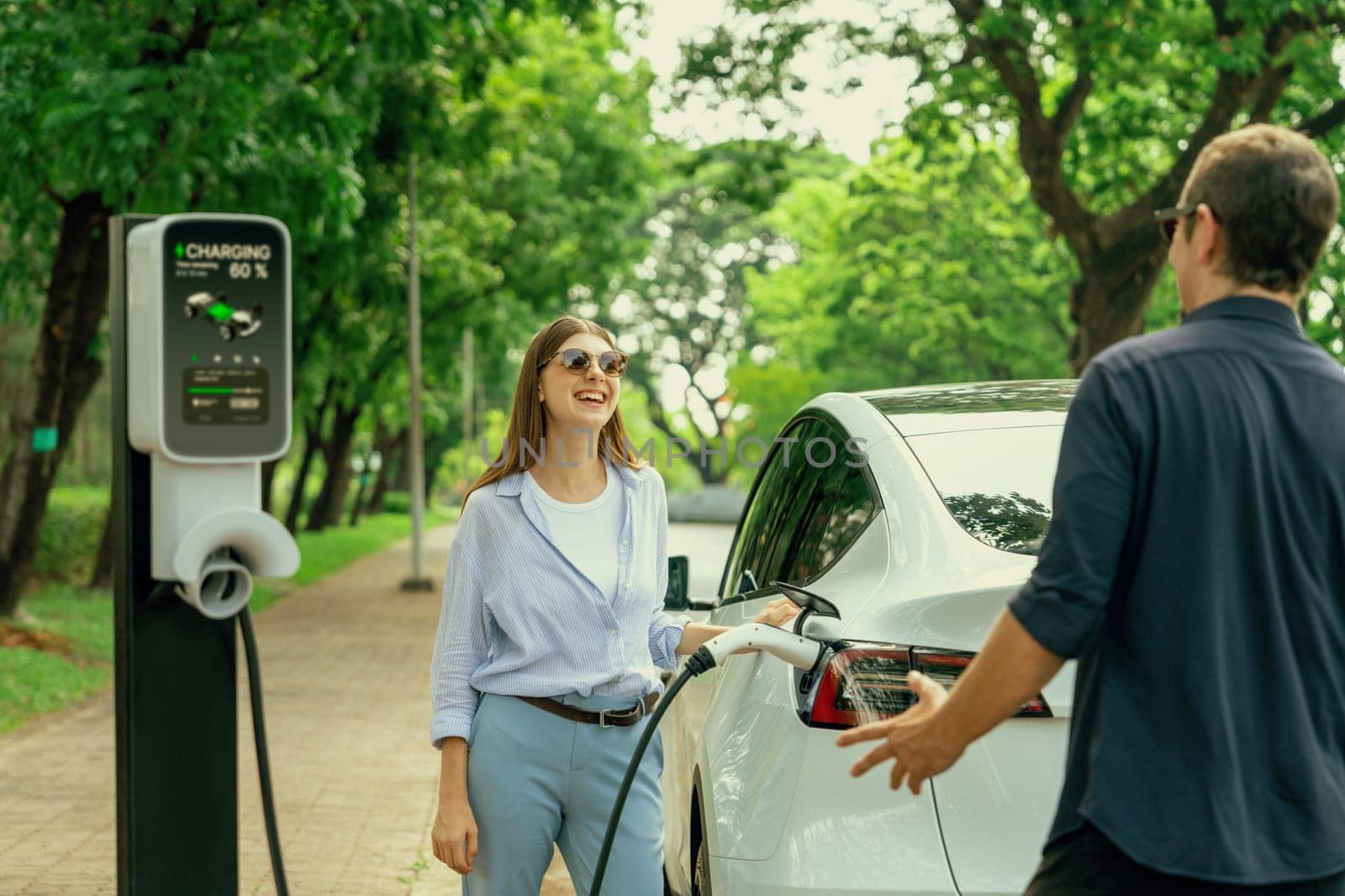 Lovely young couple wearing sun glasses recharging battery for electric car during road trip travel EV car in natural forest or national park. Eco friendly travel during vacation and holiday. Exalt