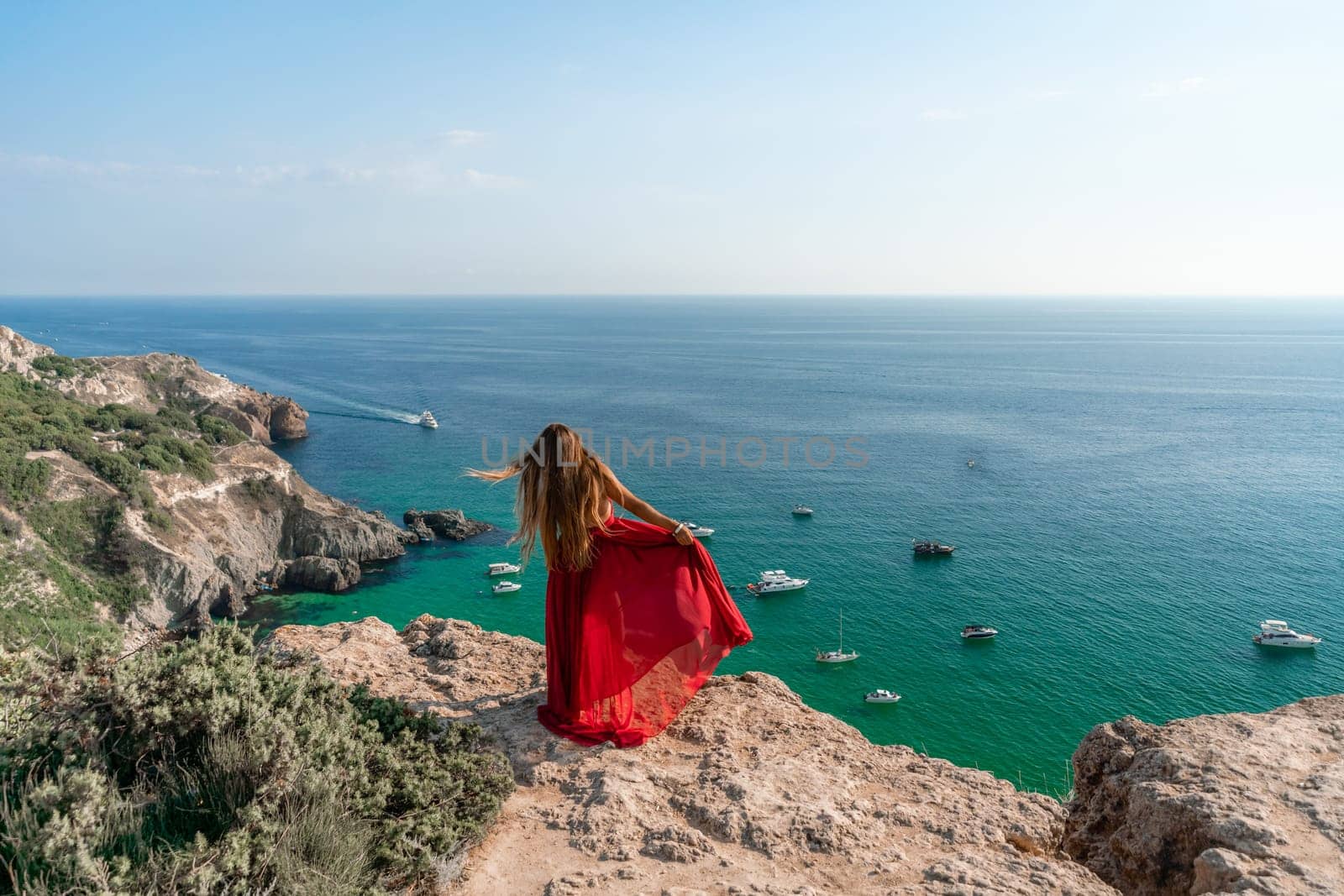 Woman sea red dress yachts. A beautiful woman in a red dress poses on a cliff overlooking the sea on a sunny day. Boats and yachts dot the background