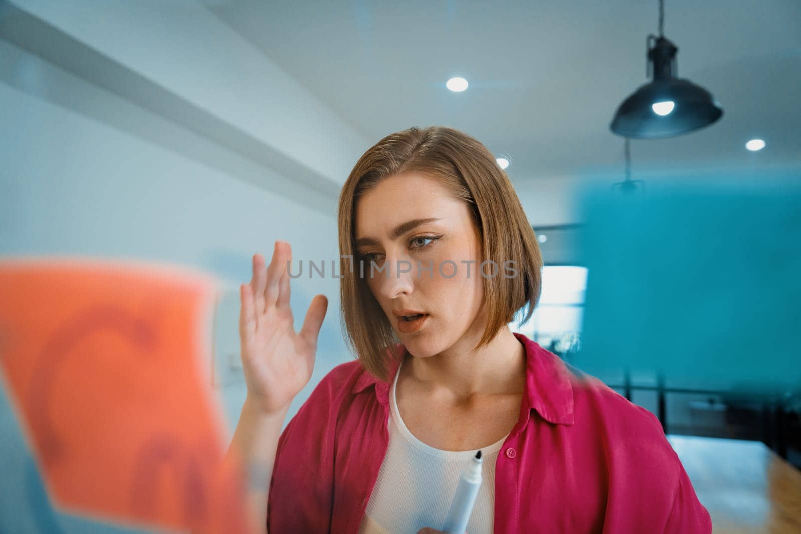 Portrait of young caucasian businesswoman thinking with confused face expression while standing in front of glass board with sticky notes and mind map at creative business meeting. Immaculate.