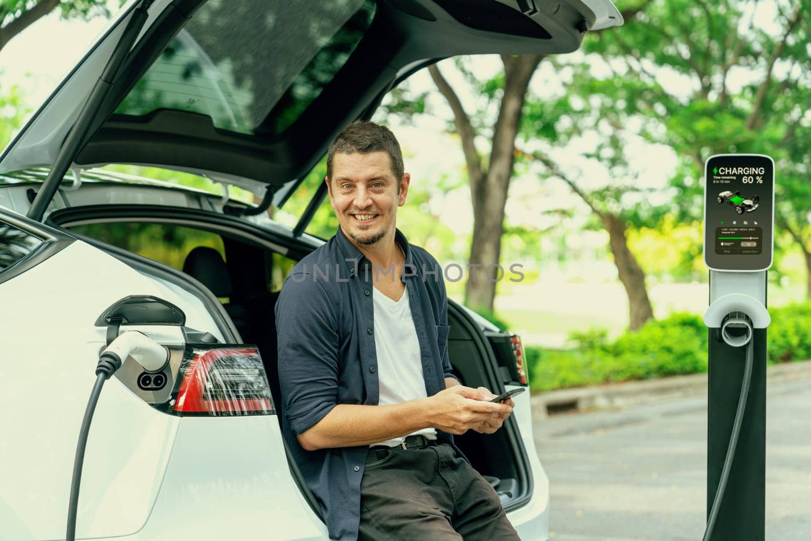 Man using smartphone online banking application to pay for electric car battery charging from EV charging station during vacation holiday road trip at national park or summer forest. Exalt