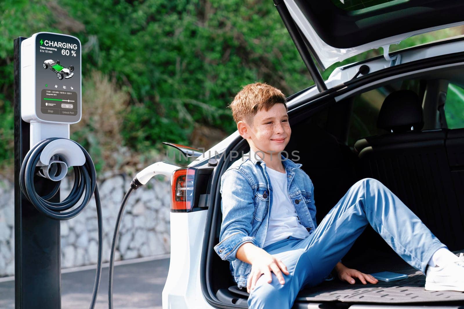 Little boy sitting on car trunk while recharging eco-friendly electric car from EV charging station. EV car road trip travel concept for alternative transportation powered sustainable energy.Perpetual