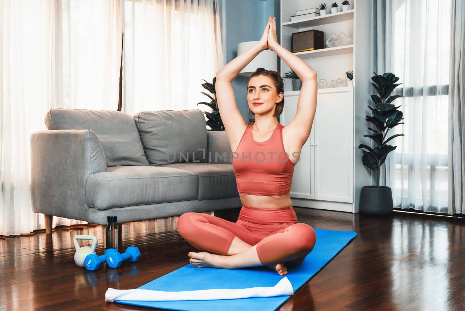 Flexible and dexterity woman in sportswear doing yoga position in meditation posture on exercising mat at home. Healthy gaiety home yoga lifestyle with peaceful mind and serenity.