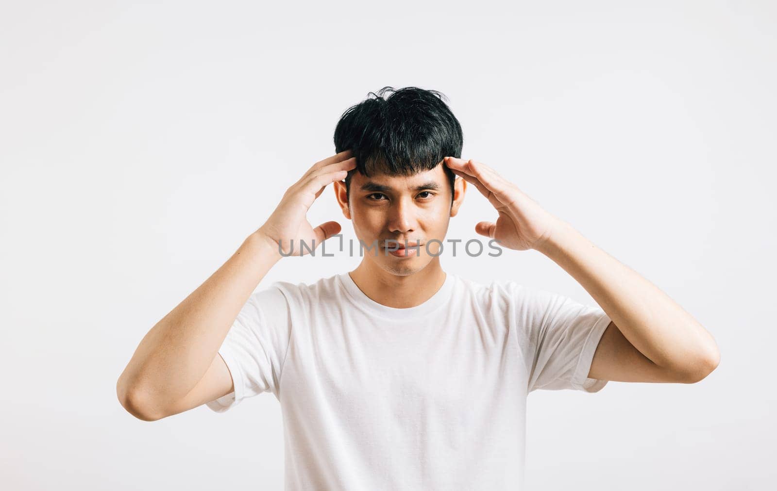 A portrait of an Asian young man with a sad, tired expression, holding his head in his hands due to stress and a painful headache. Studio shot isolated on a white background.