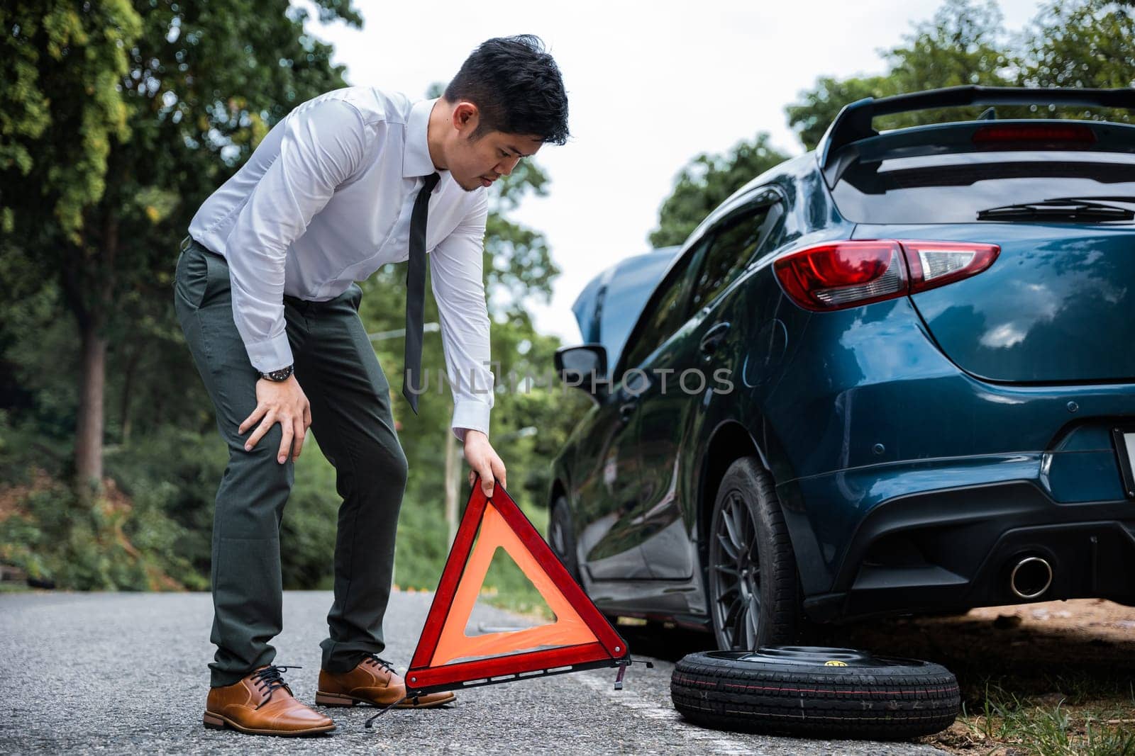 Man having car trouble and stranded at roadside with red warning triangle behind his broken car. Waiting for emergency assistance and transportation services.