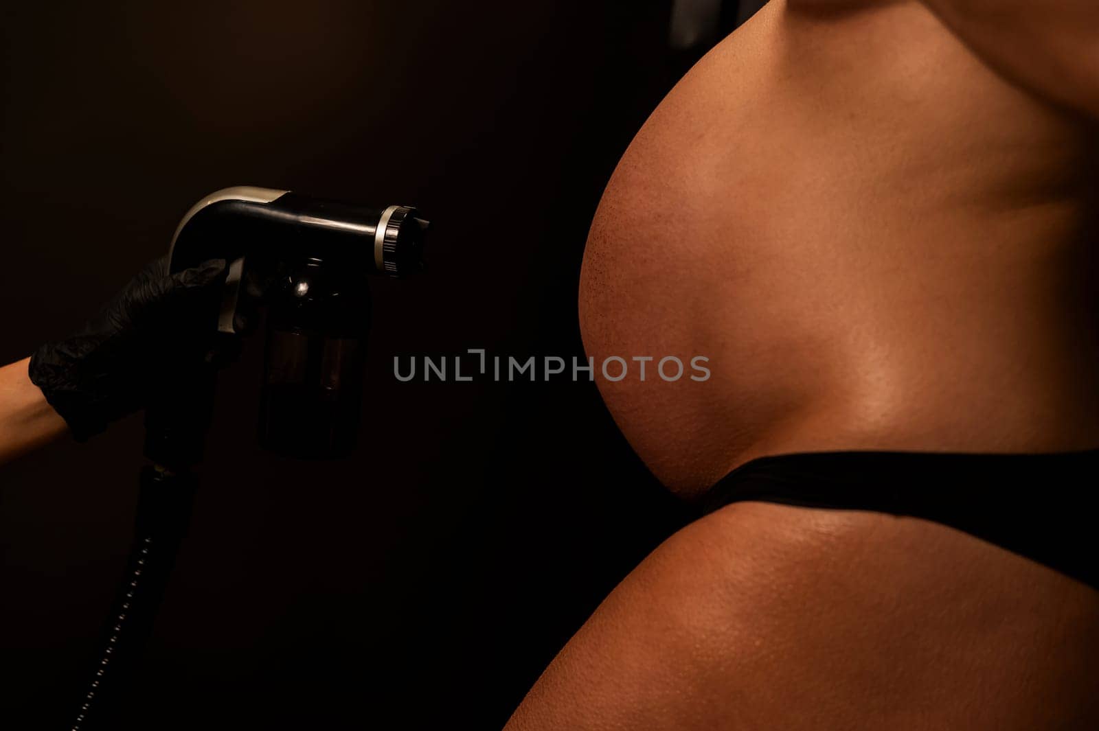 Close-up of the belly of a pregnant woman undergoing an instant tanning procedure. The master applies bronzer with a spray