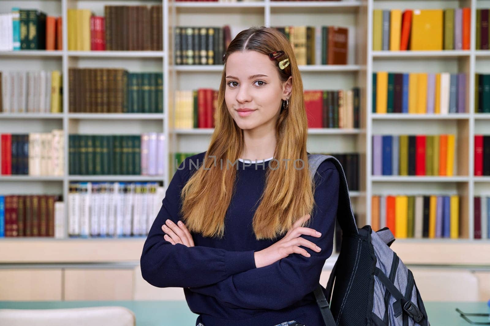 Portrait of high school student girl with backpack in library classroom. Red-haired teenage female 16, 17 years old looking at camera. Education, adolescence, school concept