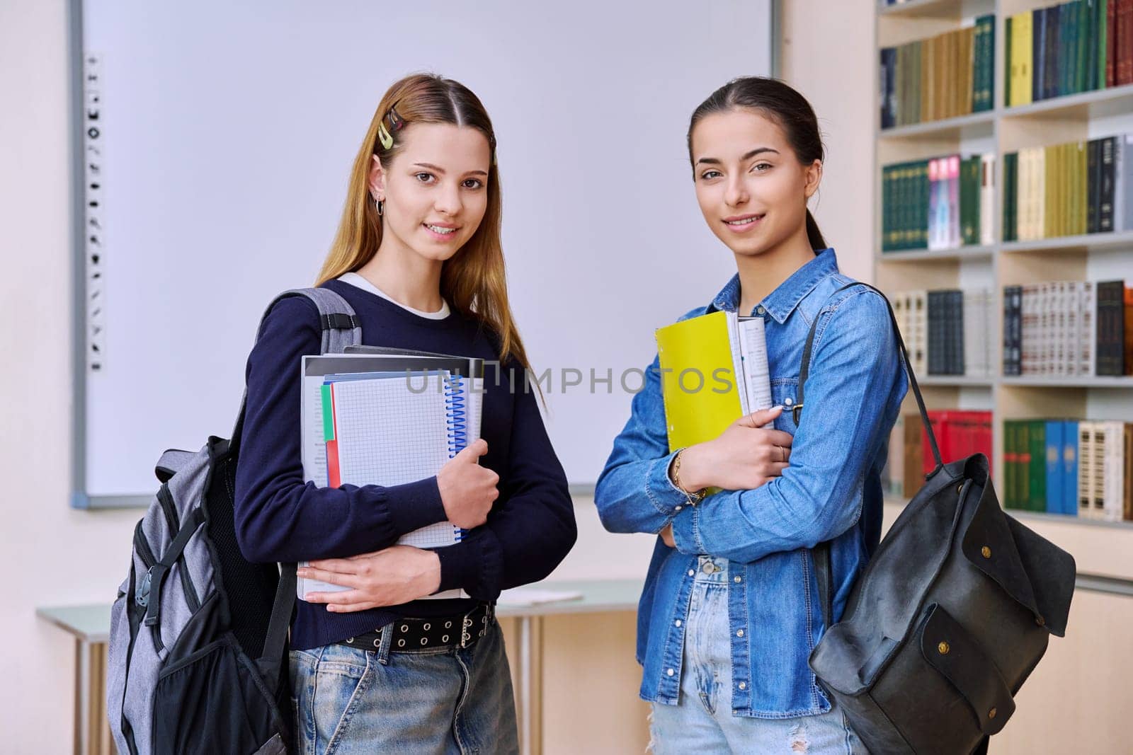Portrait of two teenage female students together in library classroom. Smiling teenage girls classmates with backpacks textbooks looking at camera. High school education adolescence friendship concept