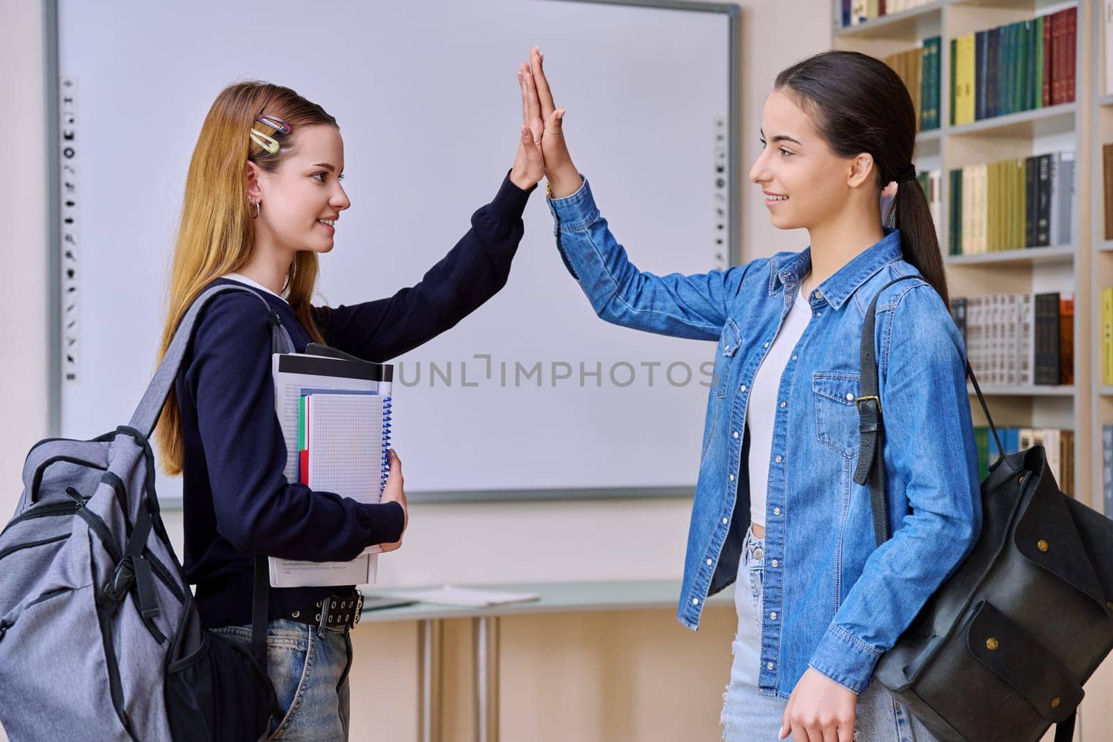 Teenage female students greeting high-fiving with hands in library class. Smiling teenagers from their classmates with textbooks and backpacks. High school education adolescence friendship communication concept