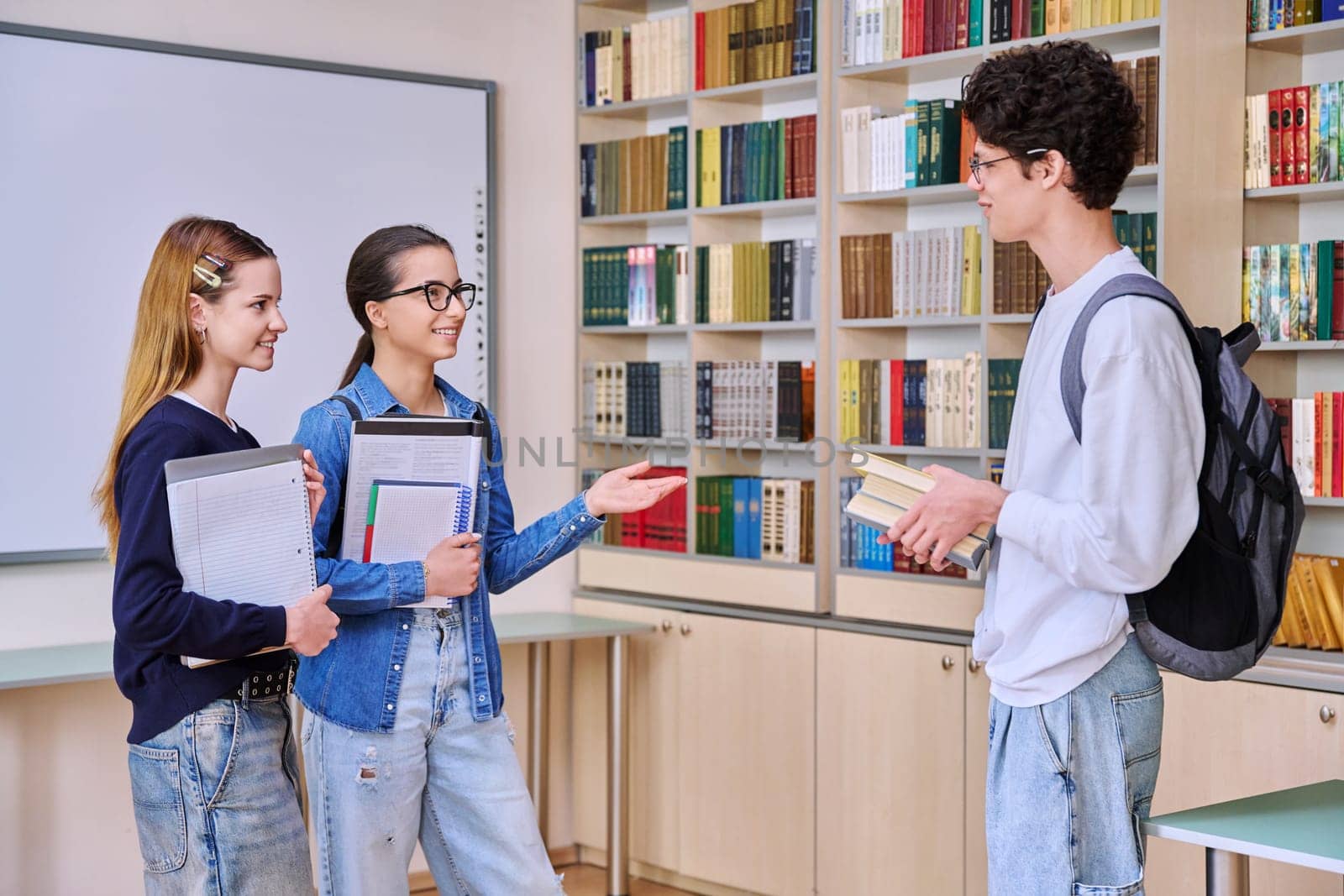 Group of teenage high school students in library classroom. Three schoolchildren 16, 17 years old, talking laughing together. Education, adolescence, friendship communication concept