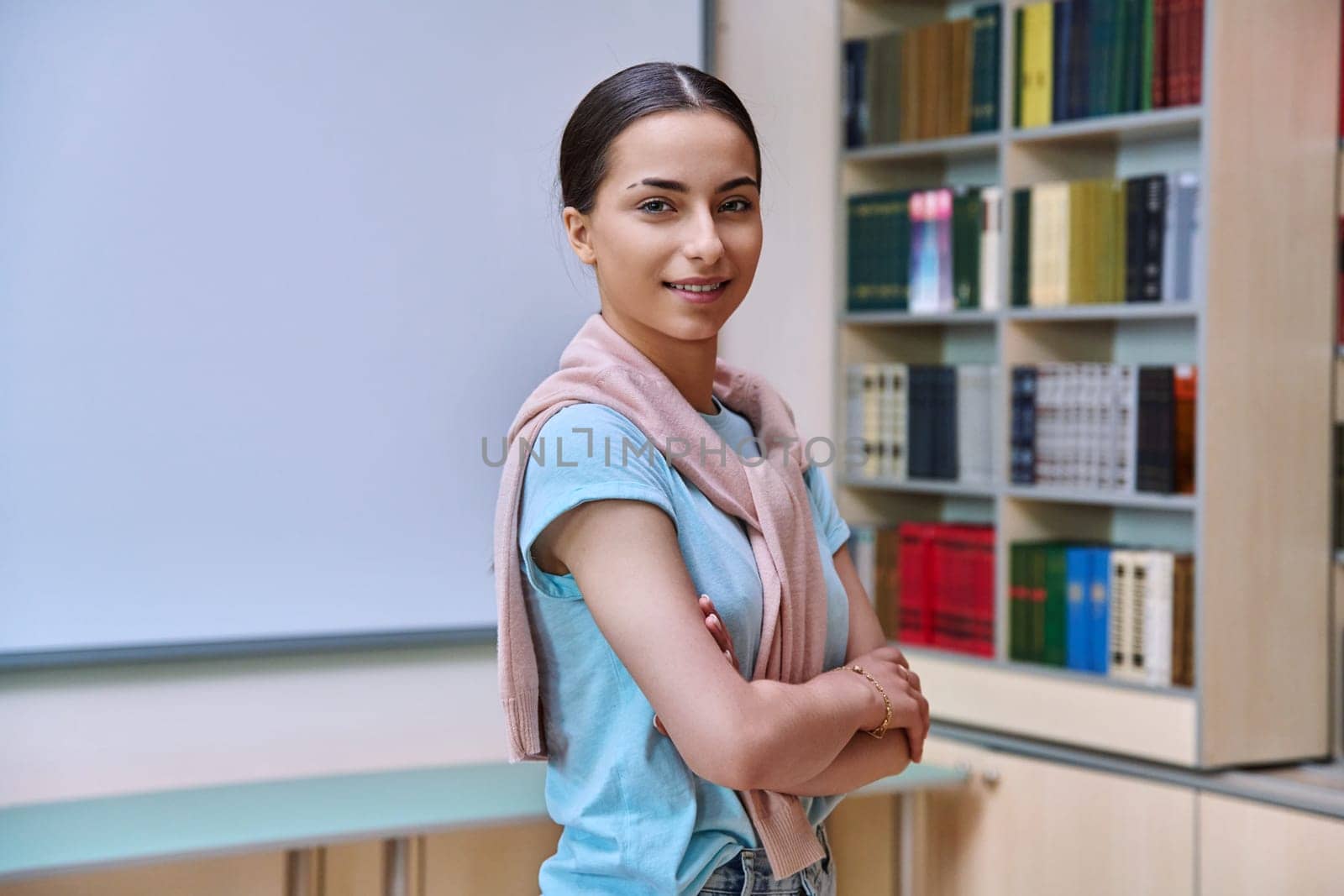 Portrait of smiling teenage girl student. Positive female teenager 16,17 years old with crossed arms inside high school building, background of library shelves books. Education, adolescence, lifestyle