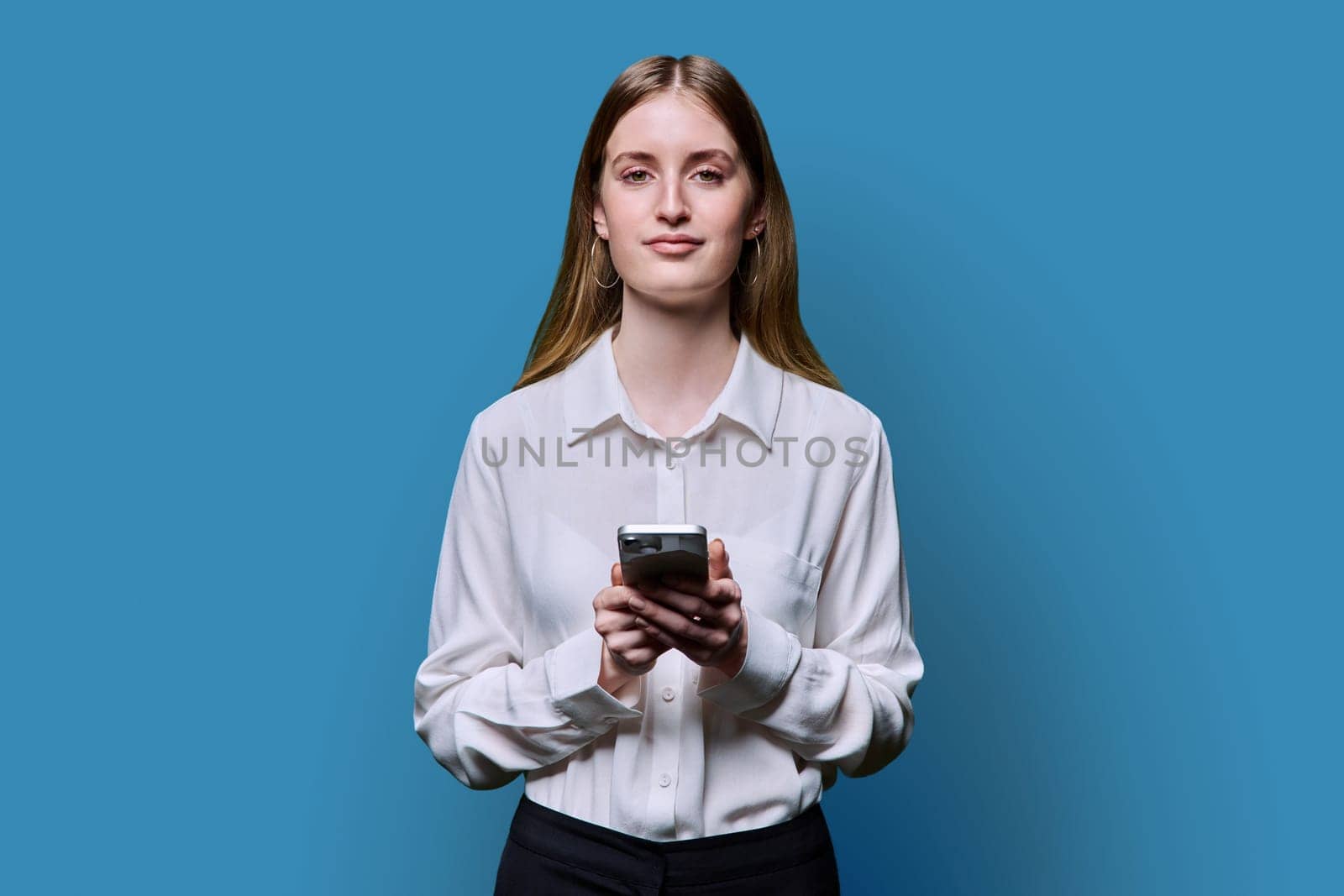 Teenage student girl 16, 17, 18 years old in white shirt holding smartphone in hands, smiling looking at camera on blue studio background. Education, technology, high school, college, lifestyle, youth concept