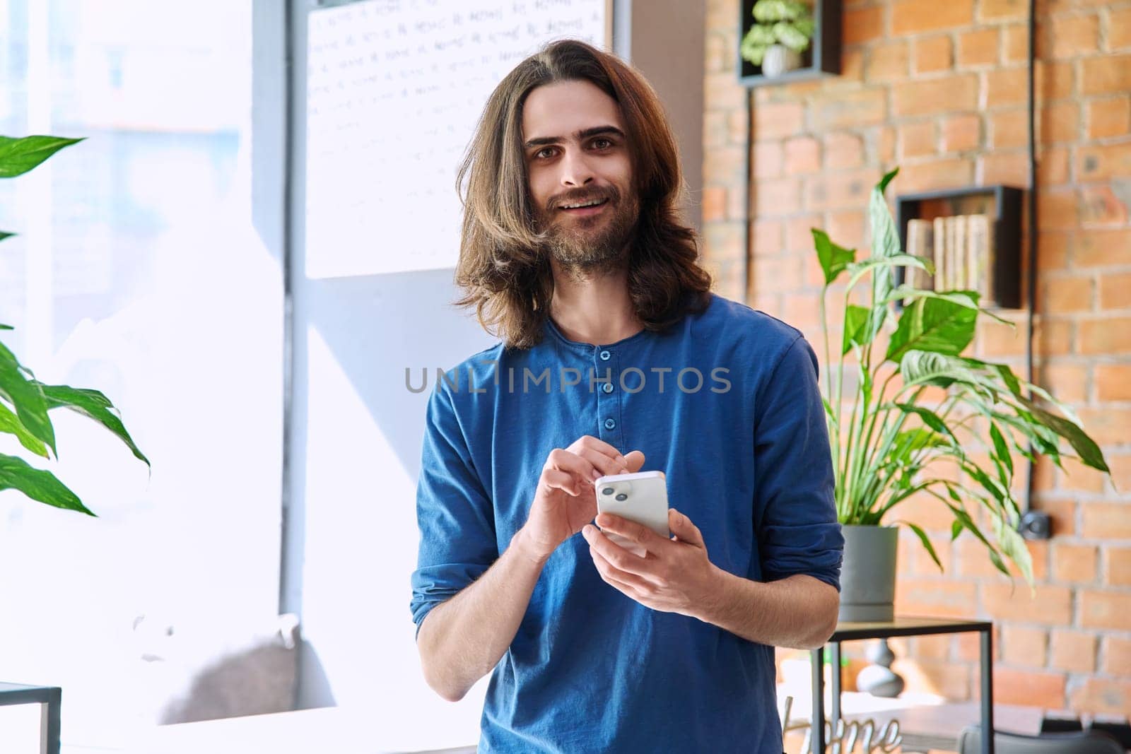 Young 30s stylish handsome bearded man with long hair using smartphone smiling guy looking at camera. Mobile technologies network Internet applications for leisure work business communication education