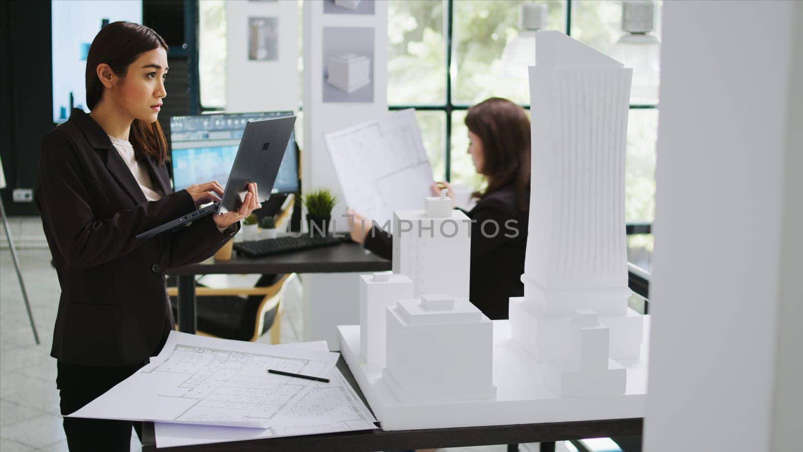 Asian contractor takes notes about 3d printed model on laptop, examining outline and scale of building maquette in architectural agency office. Cad specialist measuring layout for new project.