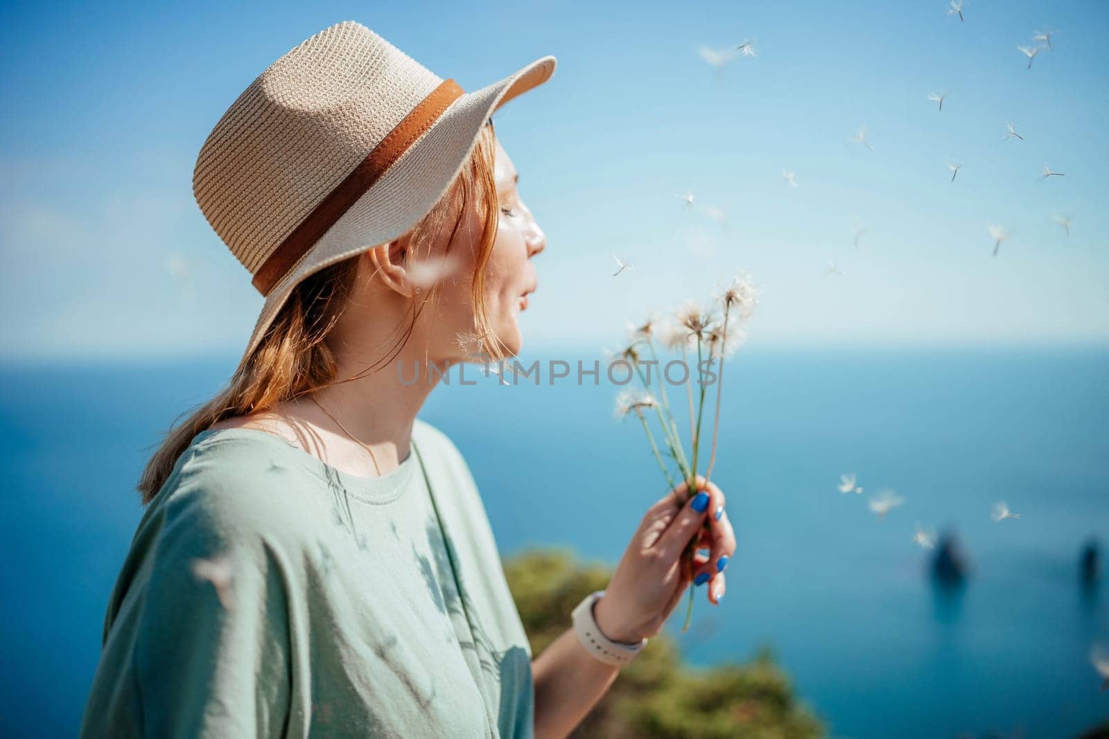 A woman wearing a straw hat is blowing on a bunch of dandelions. The scene is set on a beach with a blue ocean in the background. The woman is enjoying the moment and the beauty of the flowers