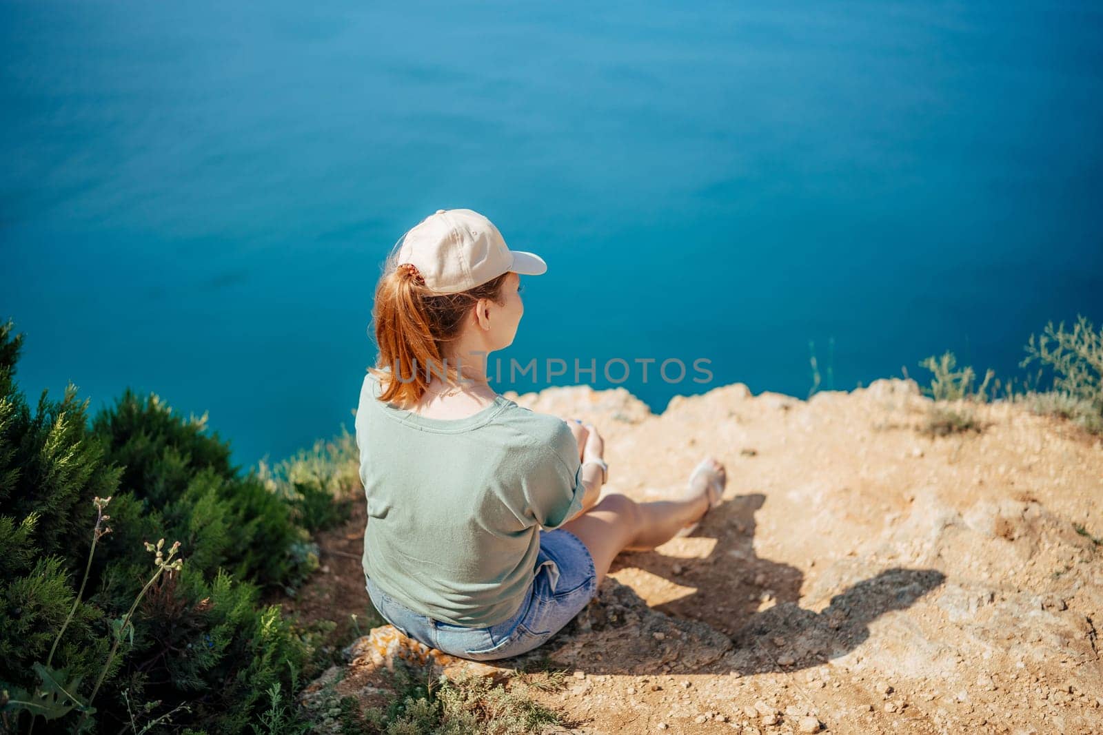 A woman is sitting on a rock overlooking the ocean. She is wearing a hat and a green shirt