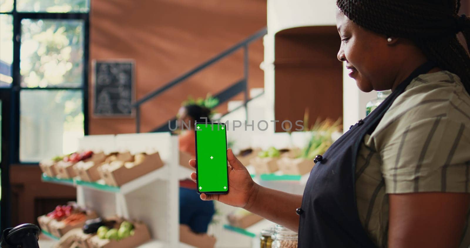 African american vendor holding phone with greenscreen, presenting isolated chromakey layout on device at farmers market checkout. Woman using copyspace screen on smartphone. Tripod shot.