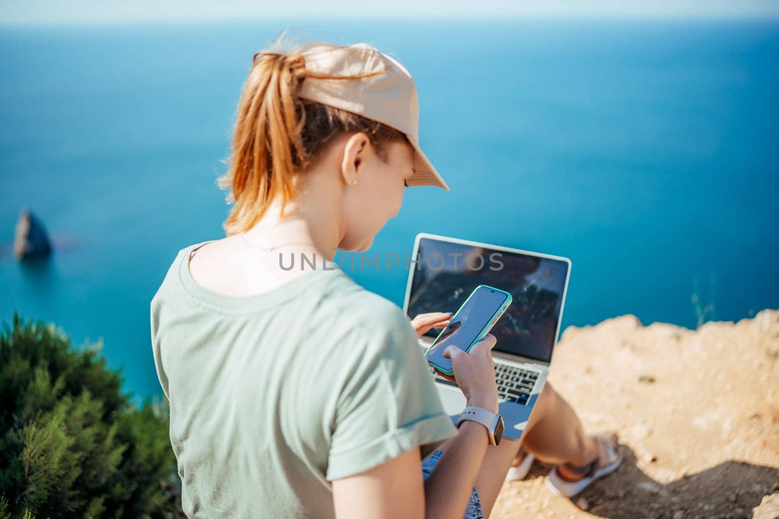 A woman is sitting on a rock by the ocean, using her laptop and cell phone. She is wearing a hat and has her hair in a ponytail. Concept of relaxation and leisure