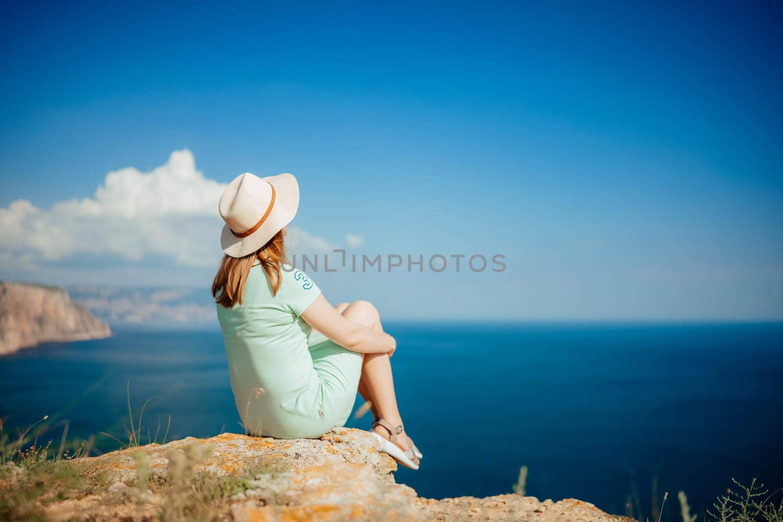 A woman is sitting on a rock overlooking the ocean. She is wearing a straw hat and a green dress. The sky is clear and blue, and the ocean is calm. Concept of relaxation and tranquility