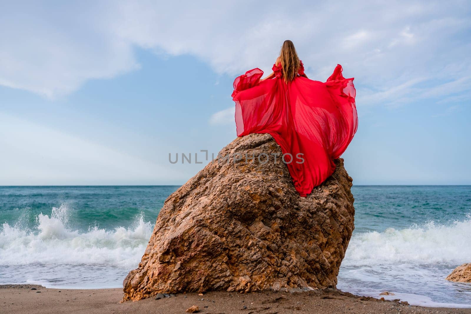 woman sea red dress. Woman with long hair on a sunny seashore in a red flowing dress, back view, silk fabric waving in the wind. Against the backdrop of the blue sky and mountains on the seashore