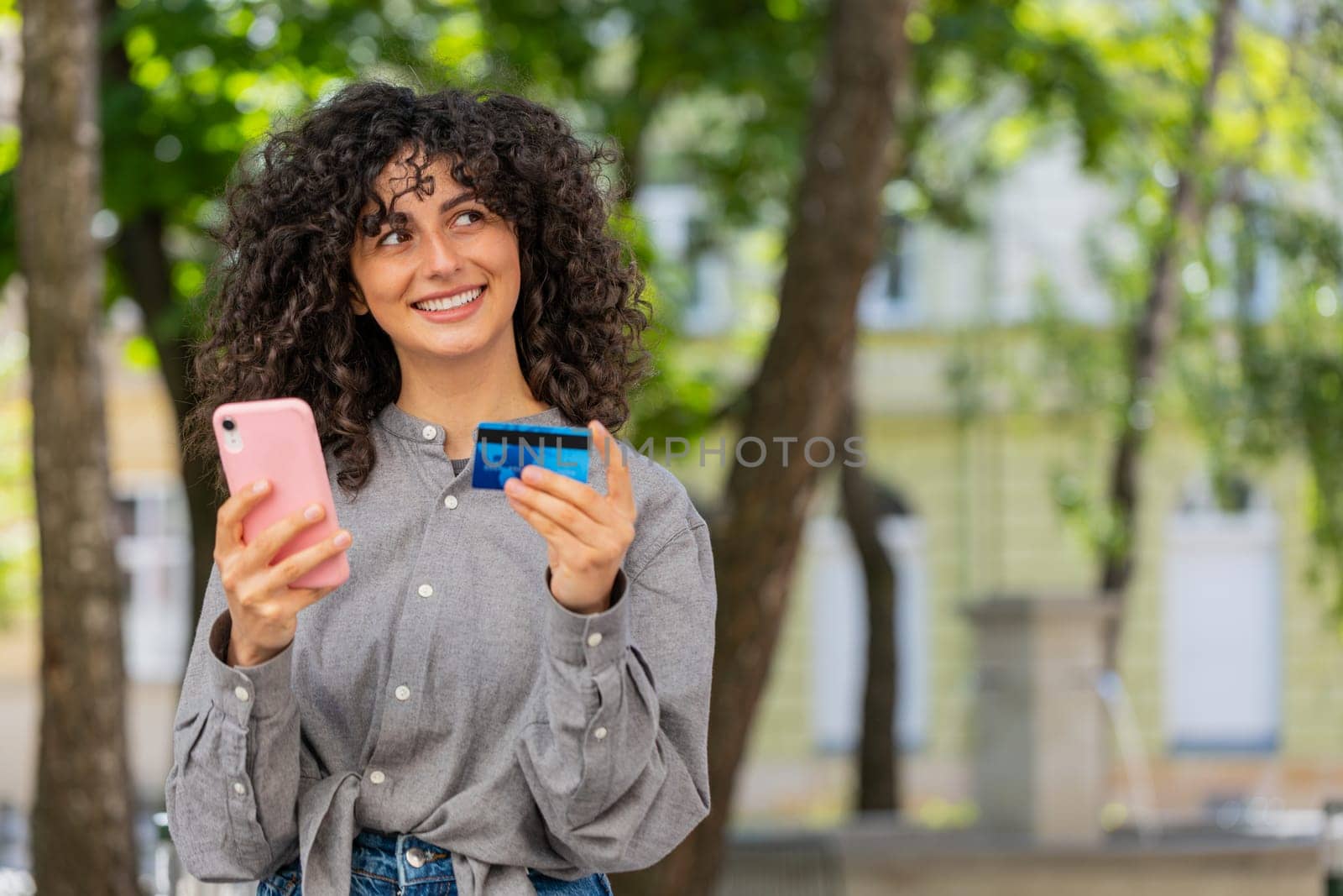 Caucasian young woman using credit bank card smartphone while transferring money, purchases online shopping, order food delivery booking hotel room outdoors. Lady girl on urban city street. Sunny park