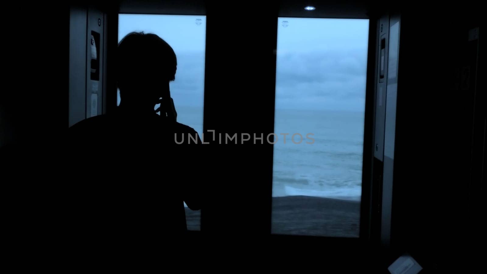 Young tourist with a smartphone looks out of the window of a moving train. Stock clip. Rear view of a male silhouette talking on phone in train. by Mediawhalestock