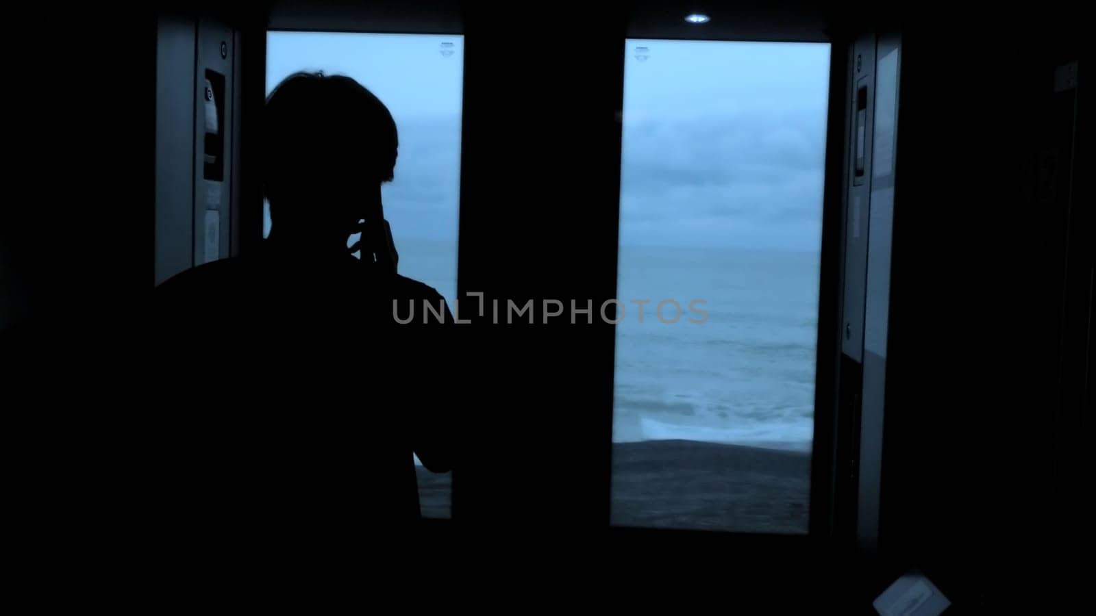 Young tourist with a smartphone looks out of the window of a moving train. Stock clip. Rear view of a male silhouette talking on phone in train