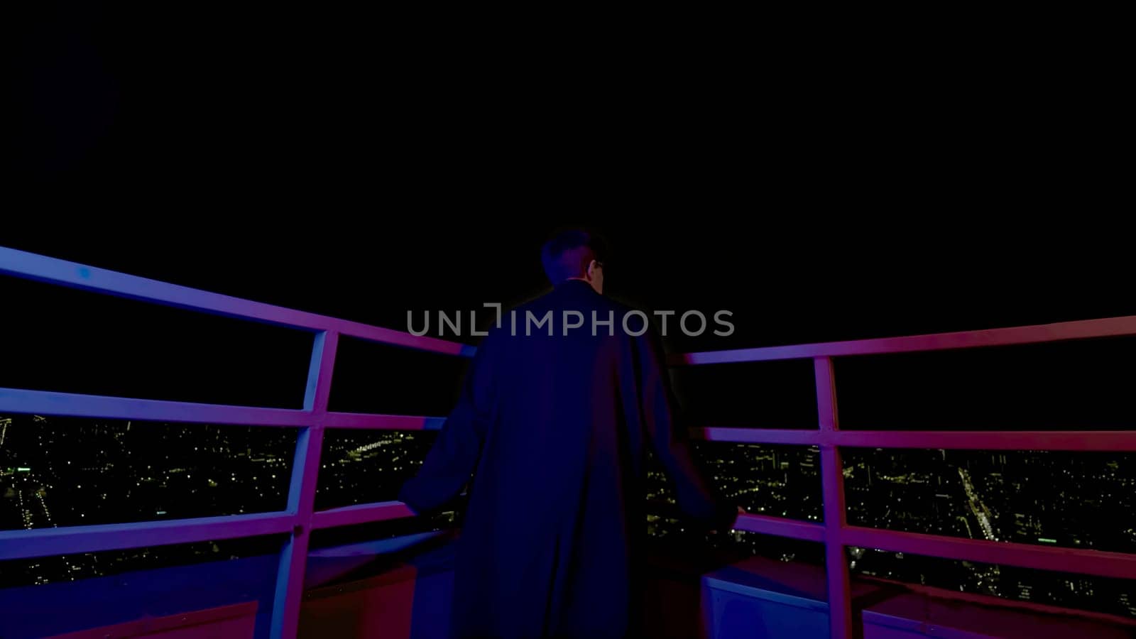 Stylish young man looks at city from roof. Media. Fashion man in coat on roof of skyscraper overlooking city. Young man looks at night city from roof of high-rise building.